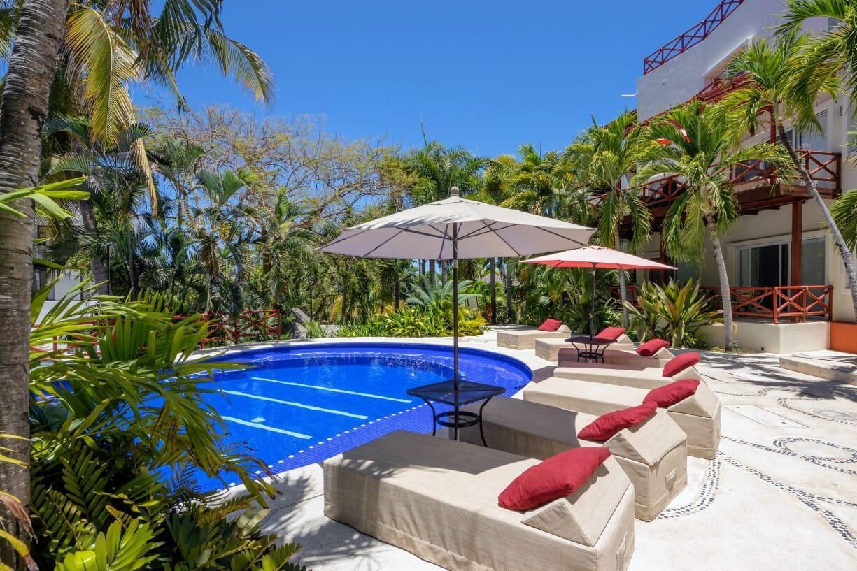Un área junto a la piscina con una piscina azul, sillas de descanso color beige con cojines rojos y una mesa con sombrilla.