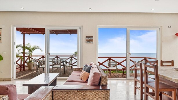 A spacious living area with a large sofa, a coffee table, and a dining table. There are multiple chairs around the dining table and a balcony with a railing. The room has large windows offering a view of the ocean.