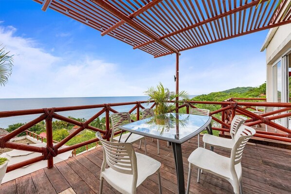 A wooden deck with a glass table and white chairs, overlooking a body of water and greenery. - Ocean View 3BR Condo, Pool, Walk to Beach at Pájaro de Fuego | Casa Vista Ballena (Sayulilla)