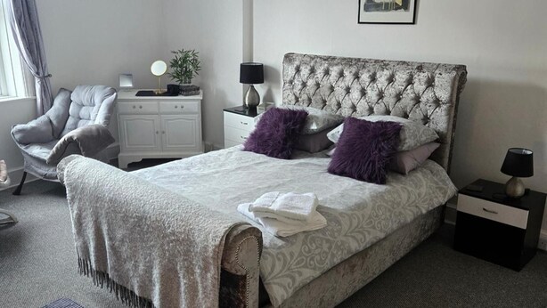 A bedroom with a tufted headboard, a bed with patterned bedding, a chair, a nightstand with a lamp, and a framed picture on the wall. - Seaview 2 Double bed Apartment 2nd Floor (Llandudno)