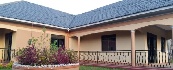 A two-story building with a tiled roof, a covered patio area, and a landscaped garden with purple foliage. - The Den Soroti Green (Kisoro)