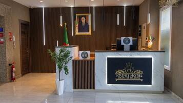 Een hotel receptie met een marmeren balie, een lijst met een portret erboven, een vlag en een brandblusser.