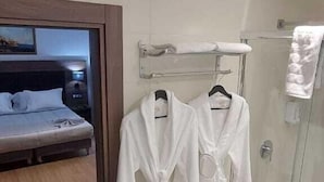 Un baño de hotel con cabina de ducha de vidrio, una cama con cabecera de madera y dos albornoces blancos colgados en un perchero.