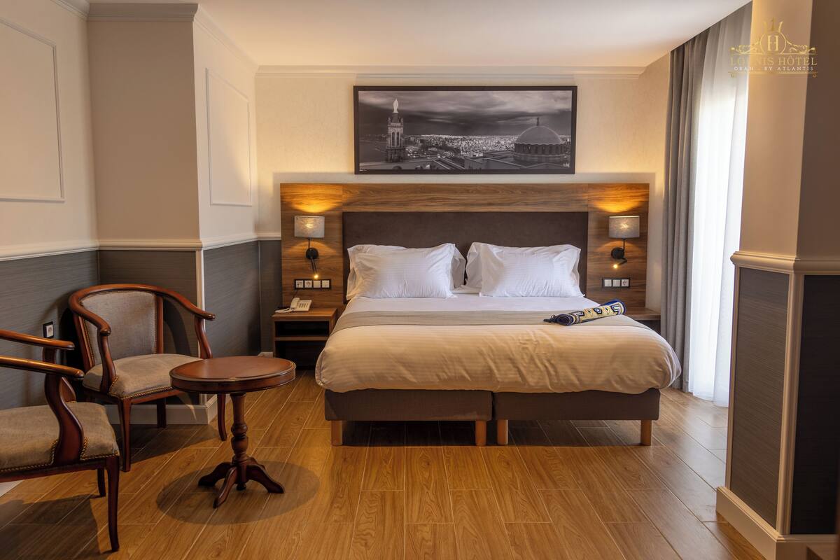 A hotel room with a large bed, two bedside tables with lamps, a chair, a round table, and a cityscape painting above the bed.