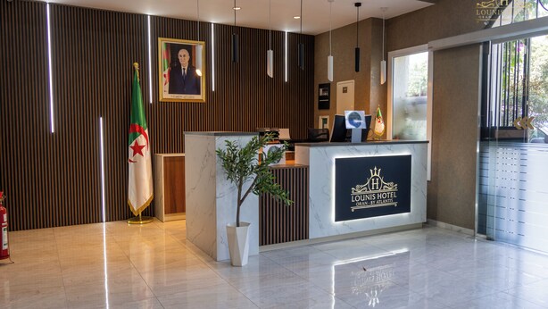 Zona de recepción de hotel con diseño moderno, que incluye un mostrador de recepción de mármol, un retrato enmarcado sobre el mostrador y una bandera a la izquierda.