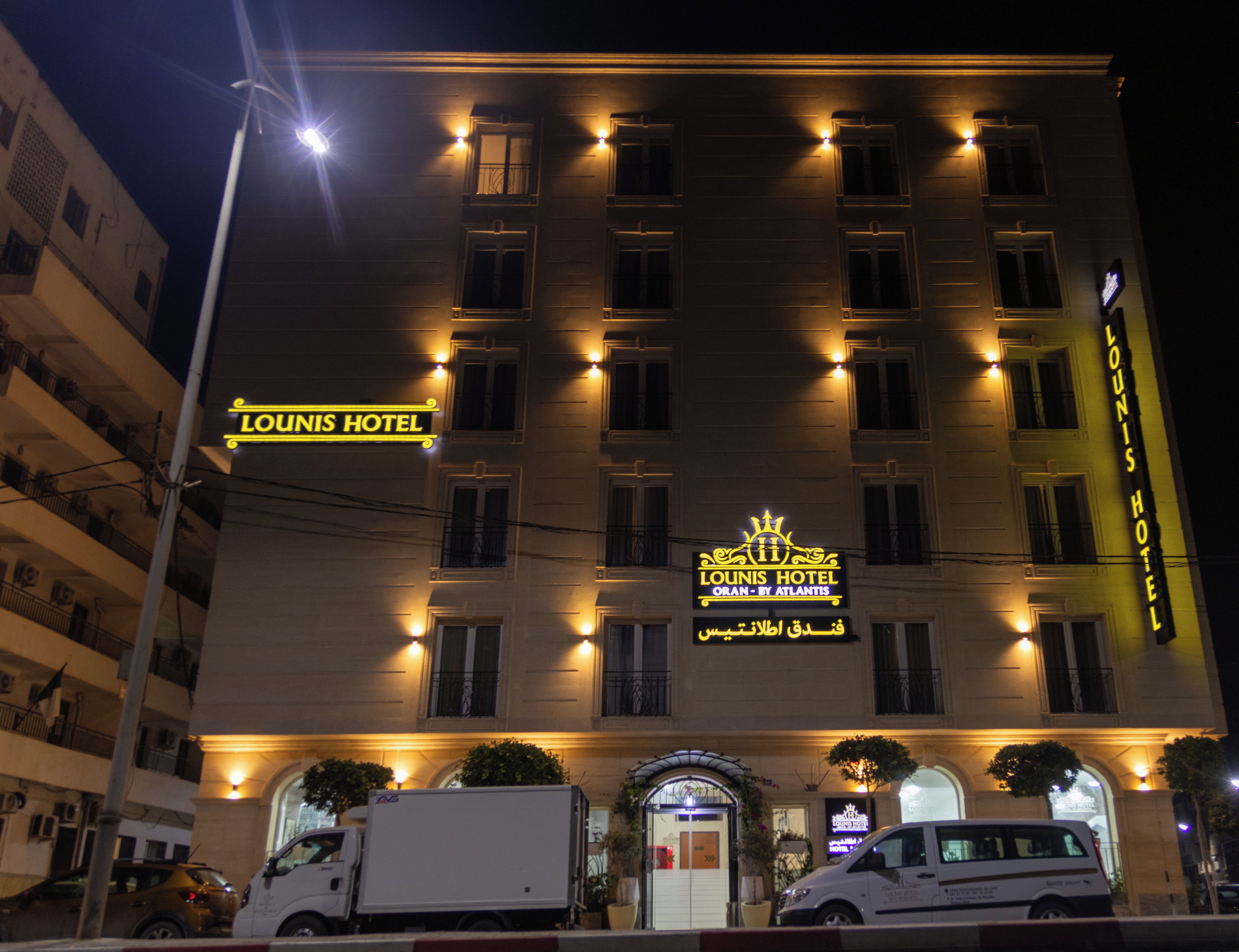 A hotel with a prominent sign reading "Lounis Hotel" and "Oran by Atlantis" in Arabic. It features a symmetrical facade with evenly spaced windows and a central entrance.