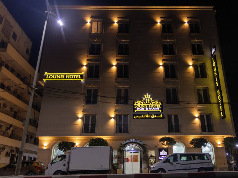 A hotel with a prominent sign reading "Lounis Hotel" and "Oran by Atlantis" in Arabic. It features a symmetrical facade with evenly spaced windows and a central entrance.