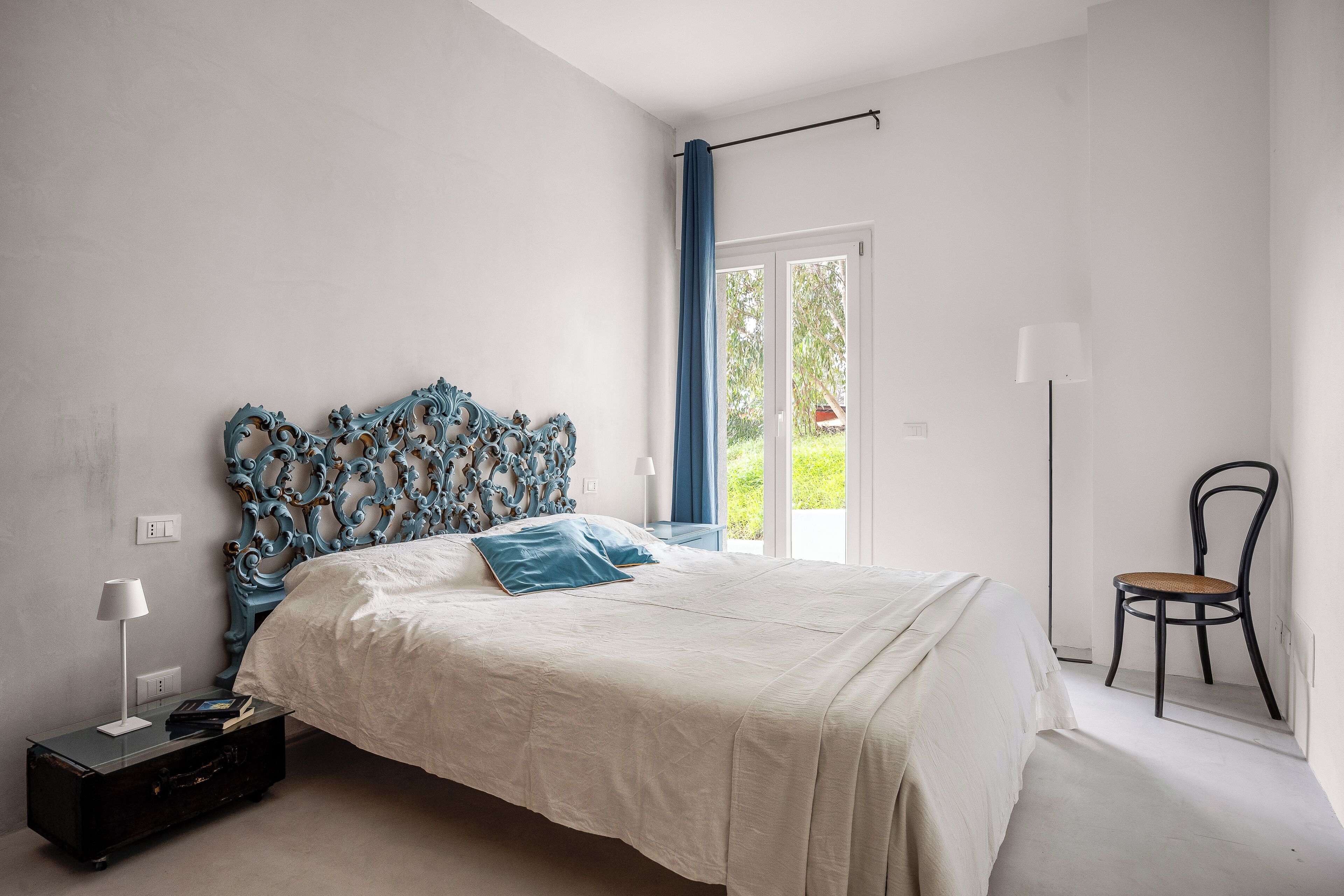 A bedroom with a large bed, a nightstand, a chair, and a window with a view of greenery.