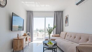 Una sala moderna con un sofá, una mesa de centro con un cuenco de fruta, un televisor y vistas al exterior a través de un ventanal grande.