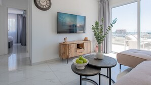 Una habitación de hotel moderna con un ventanal grande, un televisor de pantalla plana, un armario de madera, una mesa de comedor con un cuenco de fruta y un reloj en la pared.