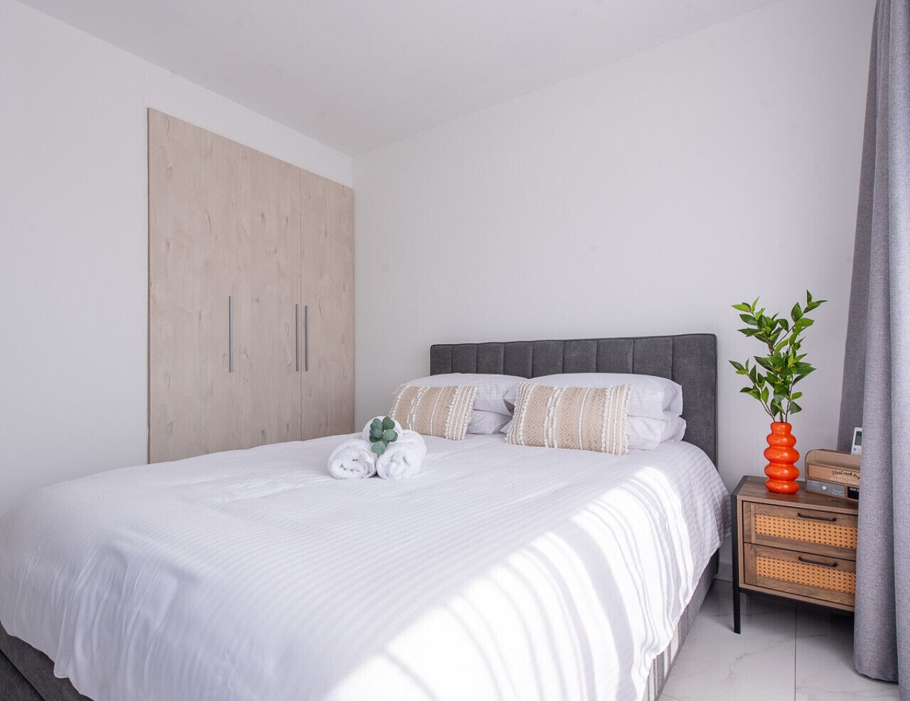 Een net opgemaakt bed met witte lakens, een grijs hoofdbord en een nachtkastje met een plant en een radio.