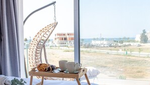 Una cama con una bandeja de madera que sostiene un croissant, un muffin y dos tazas, con vista a una playa y edificios a través de un ventanal grande.