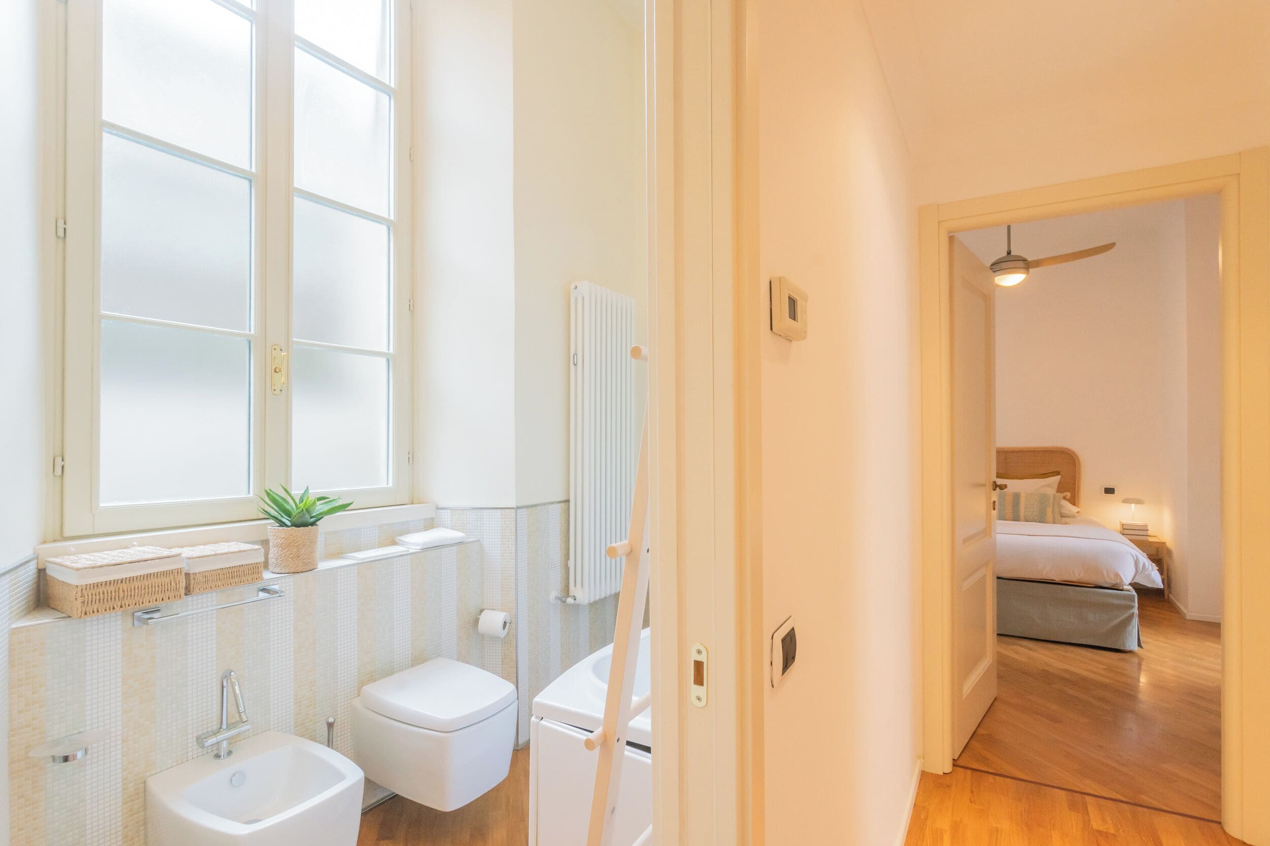 Ein modernes Badezimmer mit Badewanne, Toilette und Waschbecken, einem großen Fenster und Blick in ein Schlafzimmer mit Bett und Nachttisch.