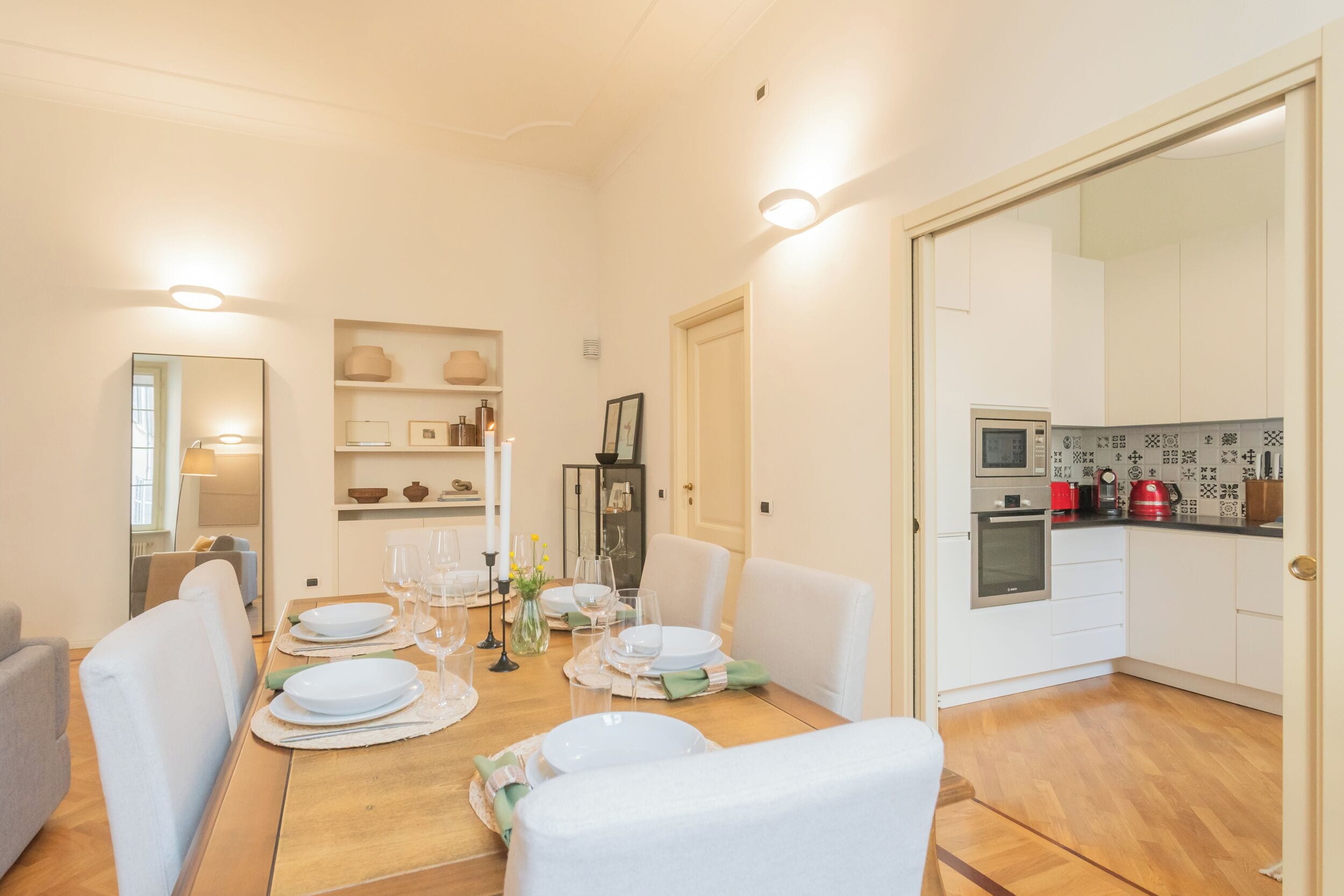 Zona pranzo con tavolo di legno apparecchiato per quattro, sedie bianche e una cucina sullo sfondo con elettrodomestici moderni.