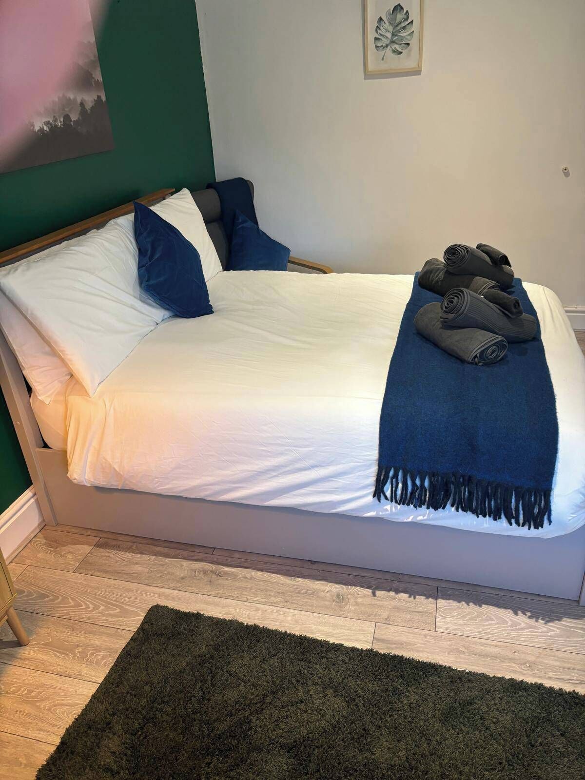 A neatly made bed with white and blue bedding, a dark blue throw blanket, and rolled-up towels on a wooden floor with a dark green rug.