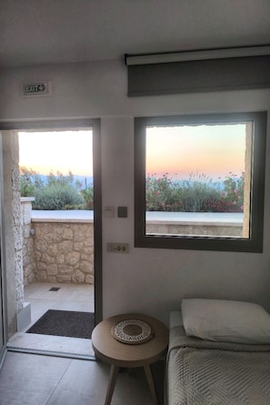 A room with a bed, a small round table, a window with a view of a garden and mountains, and an exit sign. - PALELIA living in the olive garden (Nafplio)