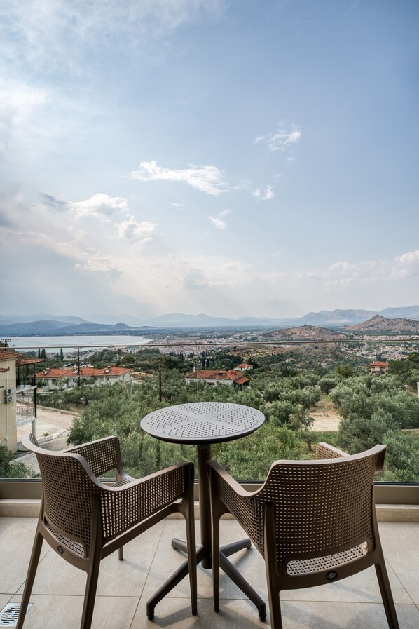 A terrace with two wicker chairs and a round table, overlooking a coastal cityscape with mountains in the distance.