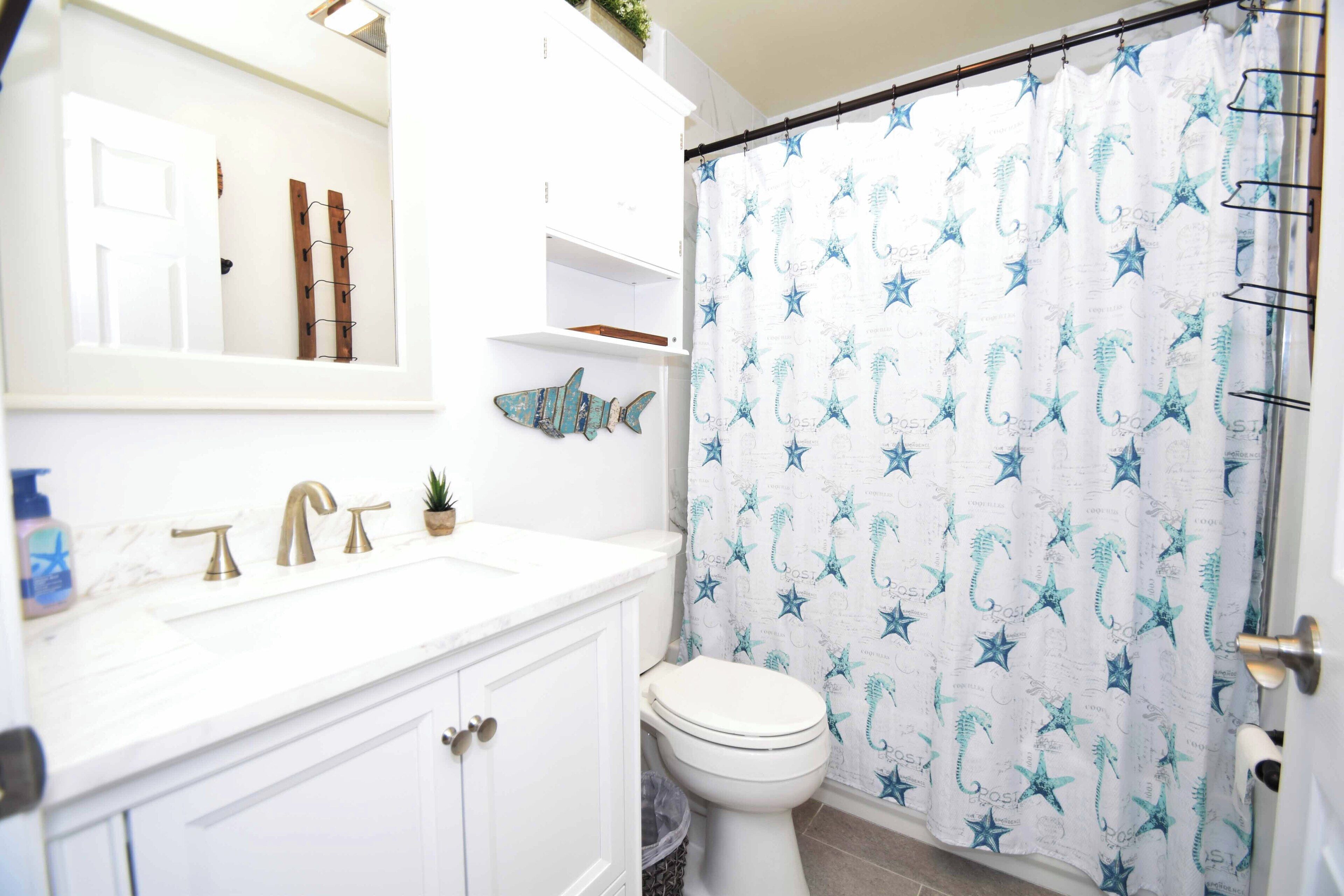Un baño con un lavabo blanco, dos grifos dorados, un inodoro blanco y una cortina de ducha con motivos de estrellas de mar y caballitos de mar.