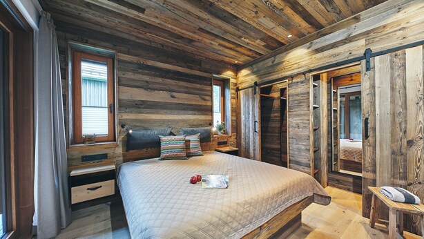 A rustic bedroom with a wooden ceiling, a bed with a quilted cover, a nightstand, and a window with curtains. - Atempause Sauerland feel-good chalet "Große Sehnsucht" (Schmallenberg)