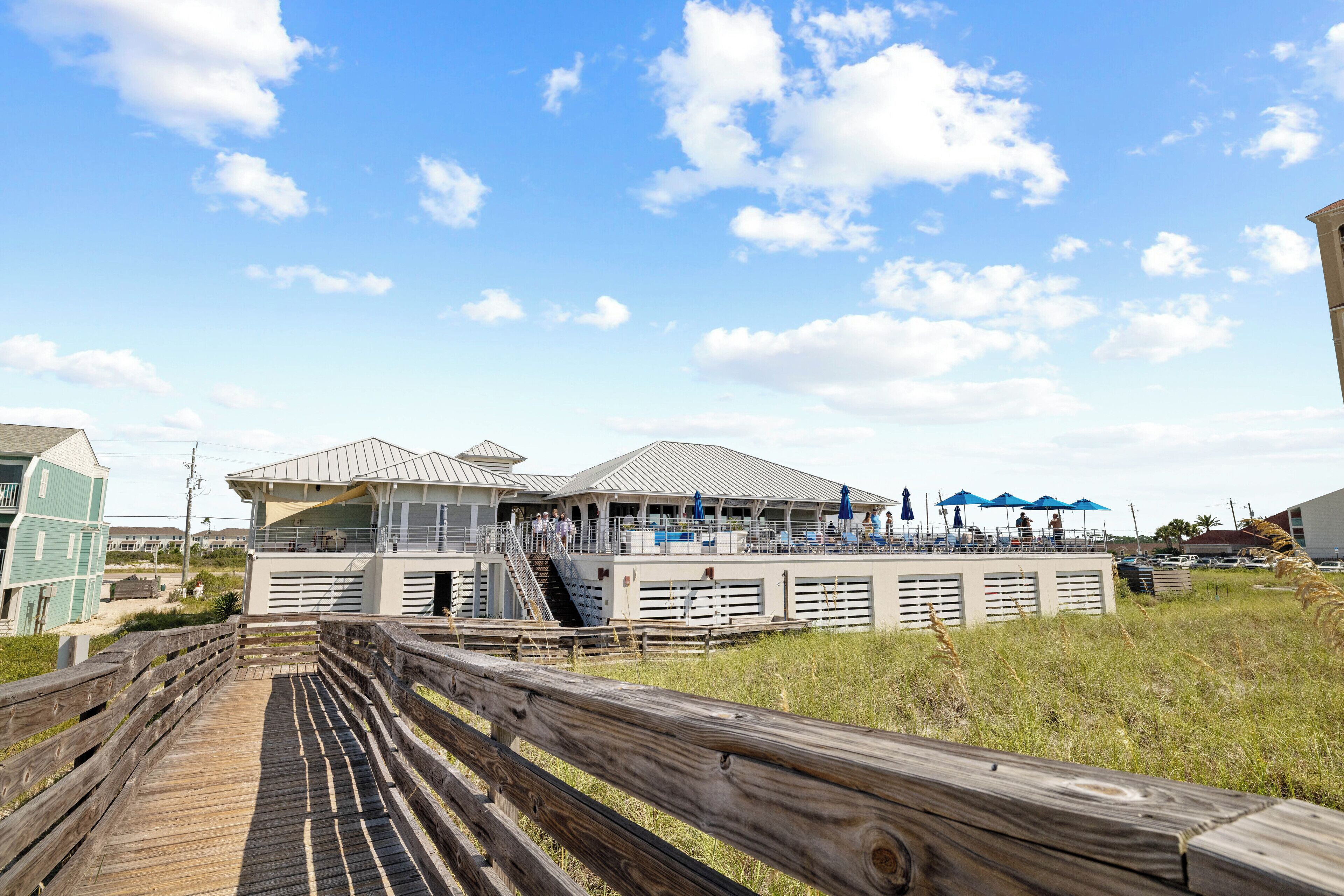 A beachfront hotel with a wooden boardwalk leading to the entrance, a covered outdoor seating area, and a view of the ocean.