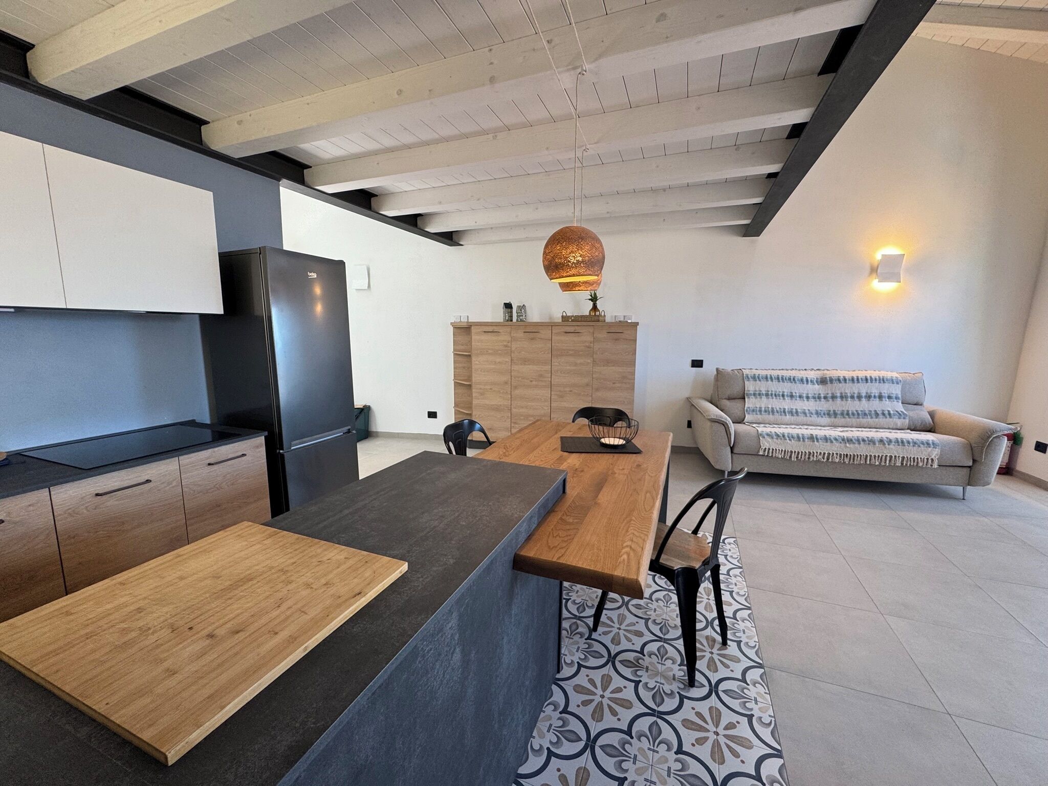 Eine moderne Küche mit einer Holzinsel, einem integrierten Kühlschrank sowie einem Essbereich mit Holztisch und -stühlen. Im Wohnbereich befindet sich eine Couch und ein Teppich mit Muster.