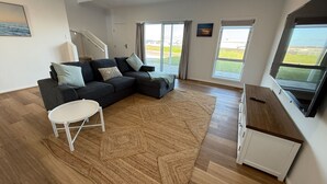 A modern living room with a sectional sofa, a white coffee table, and a large window offering a view of the outdoors. - Sea Escape - Sea Views!  Short walk to Tavern! Off Peak Special available! (Point Turton)