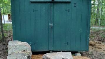 Um galpão verde com porta e o número '2' na sua fachada, rodeado por pedras e cascalho.