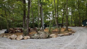 Un terrain de camping boisé avec un sentier de gravier, des chaises en bois, une pile de bois de chauffage et une petite construction.