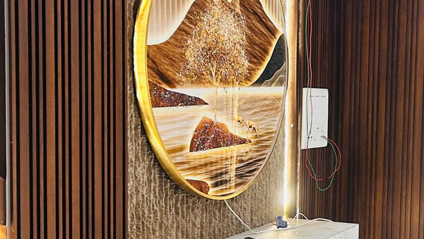 A modern bathroom with a wooden panel wall, a round mirror with a landscape design, and a built-in white shelf. - Hotel Kamakhya Inn (Chhatarpur)