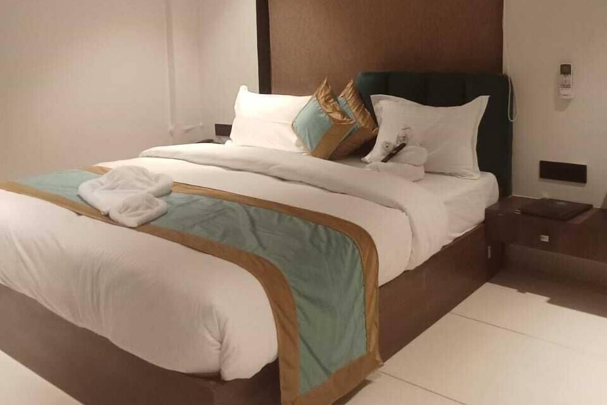 Una cama bien hecha con sábanas blancas y una manta turquesa, cabecero oscuro y mesita de noche con una lámpara.