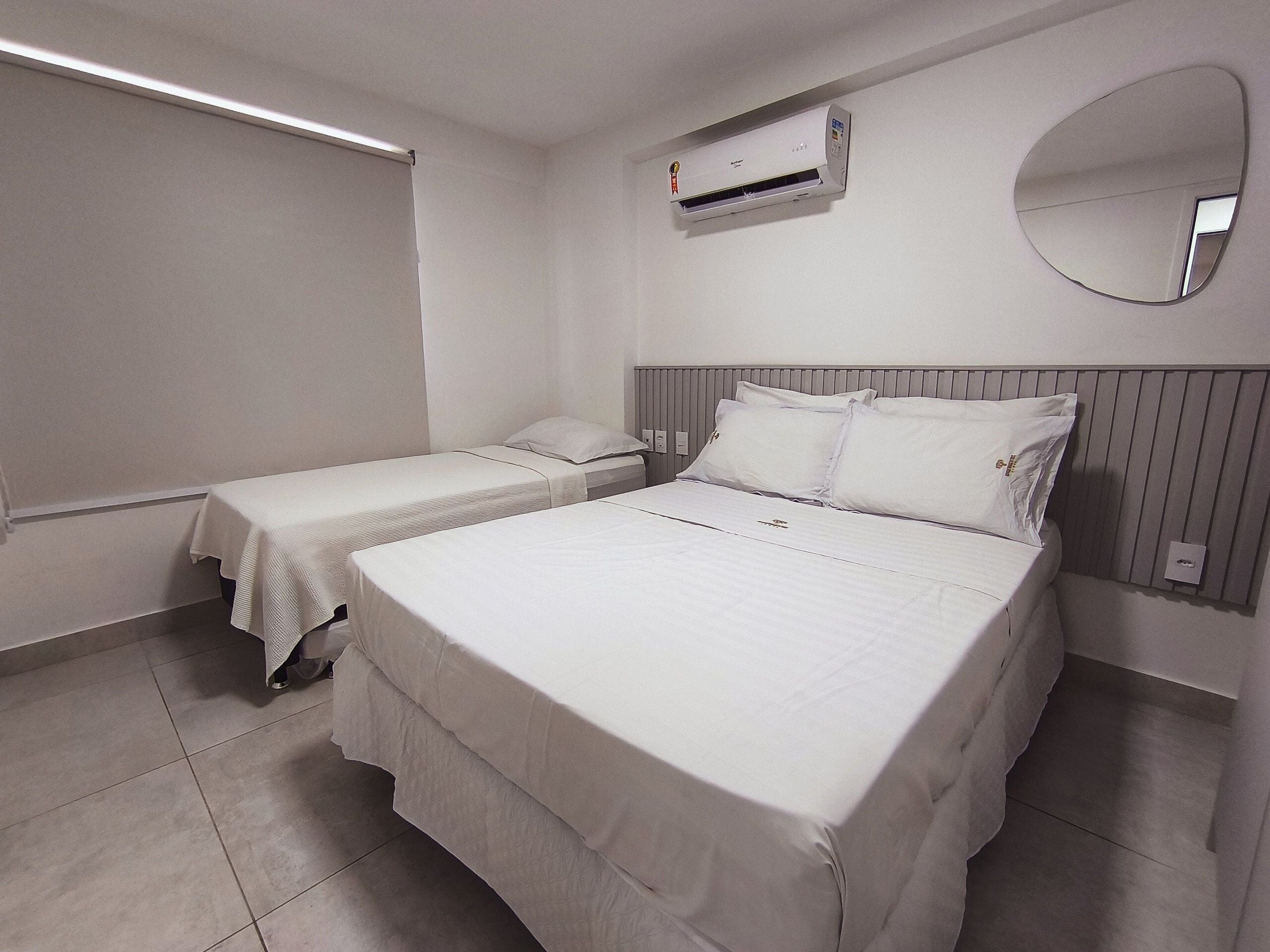 A hotel room with two single beds, a large bed, a wall-mounted air conditioner, a round mirror, and a window with blinds.