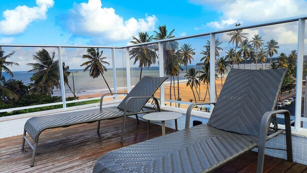 A balcony with two wicker lounge chairs and a small table, overlooking a beach with palm trees and the ocean.
