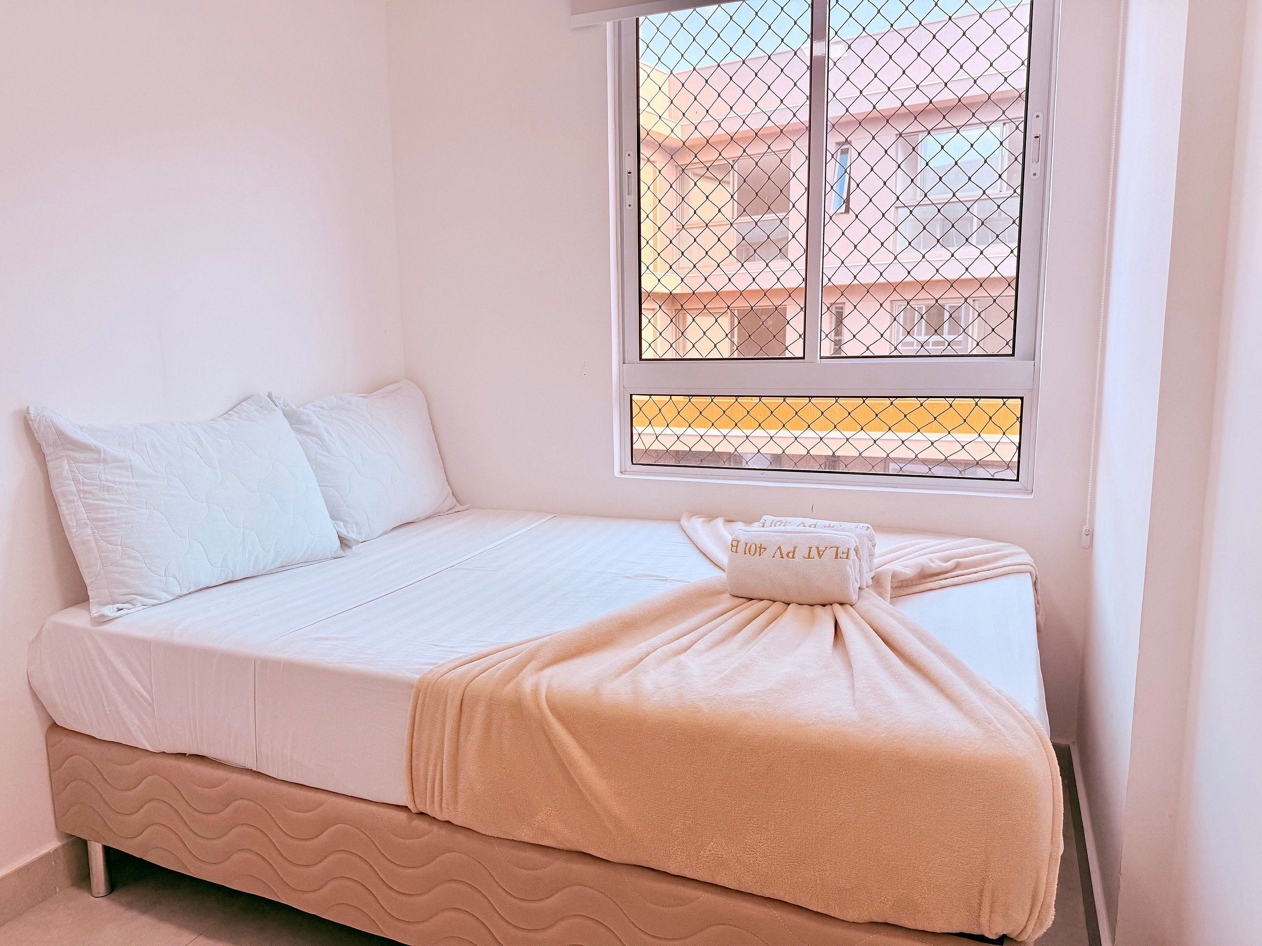Una cama individual con ropa de cama blanca, una manta color beige y una almohada. Hay una ventana con celosía metálica y una toalla sobre la cama.