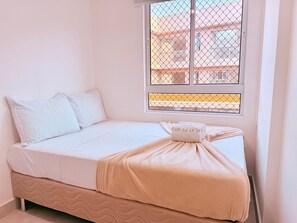 Una cama individual con ropa de cama blanca, una manta color beige y una almohada. Hay una ventana con celosía metálica y una toalla sobre la cama.