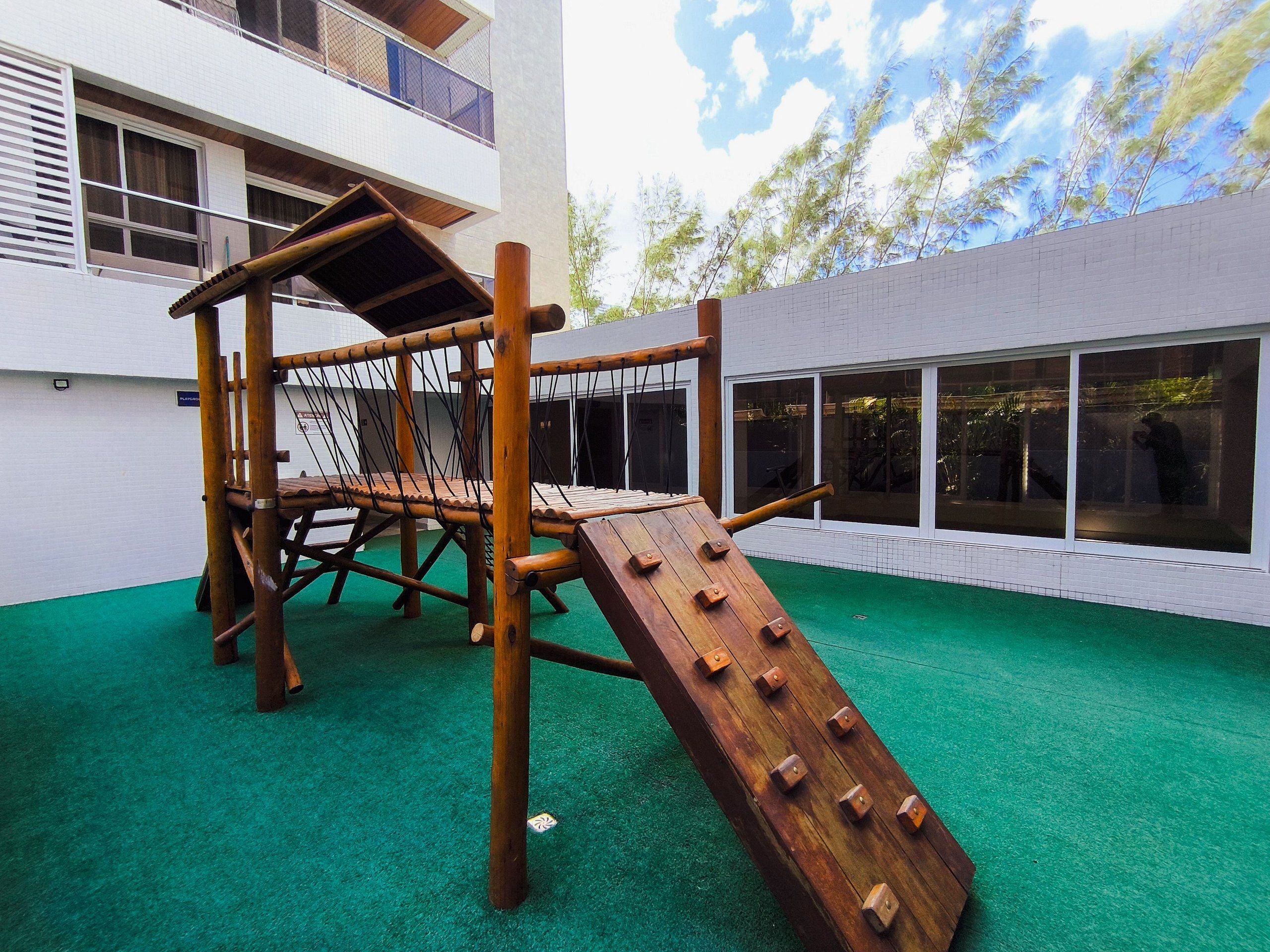 Una estructura de juego de madera en un espacio exterior con césped artificial verde, rodeada por un edificio blanco con ventanales amplios.