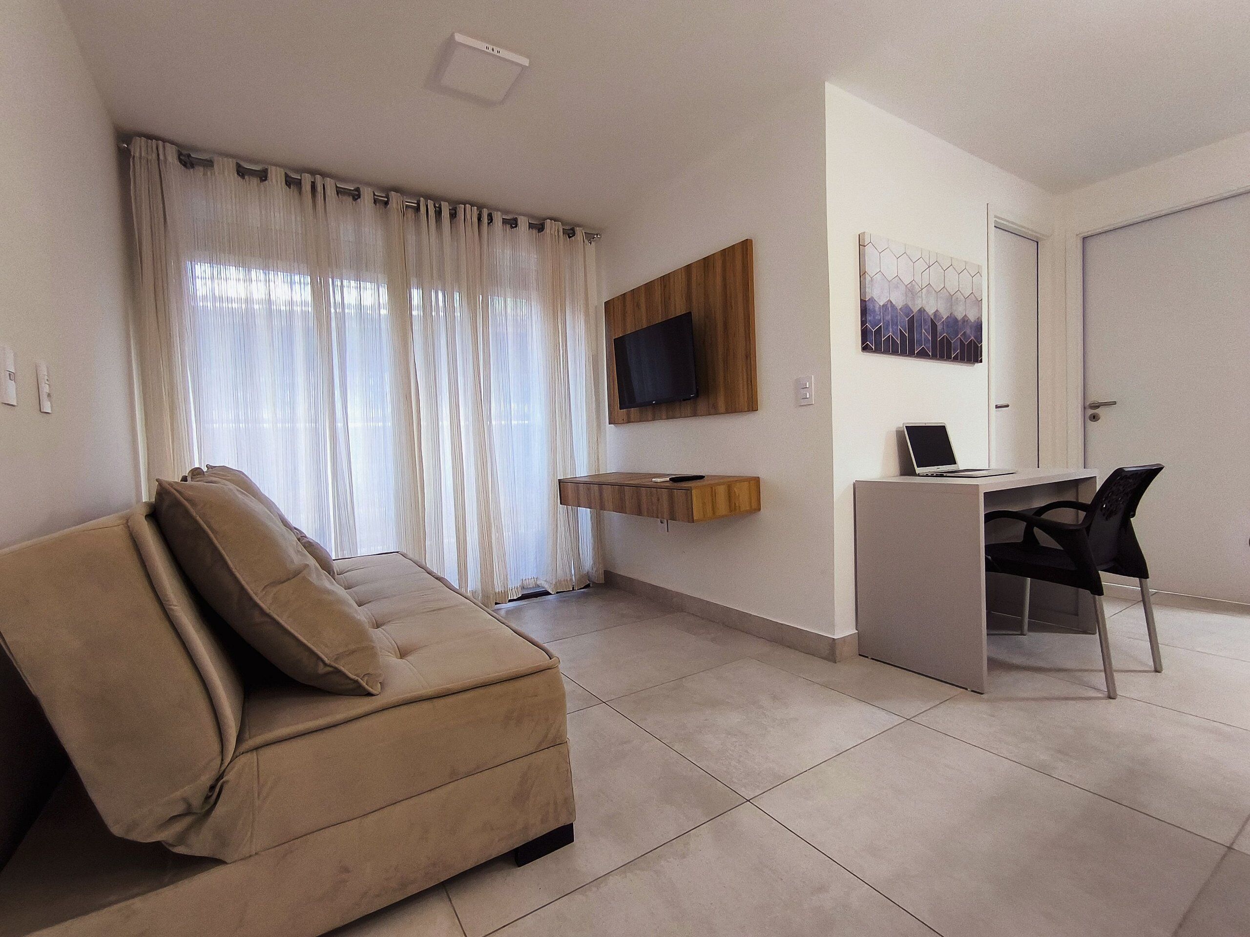 A modern living room with a beige sofa, a wooden TV stand, a desk with a laptop, and a large window with sheer curtains.