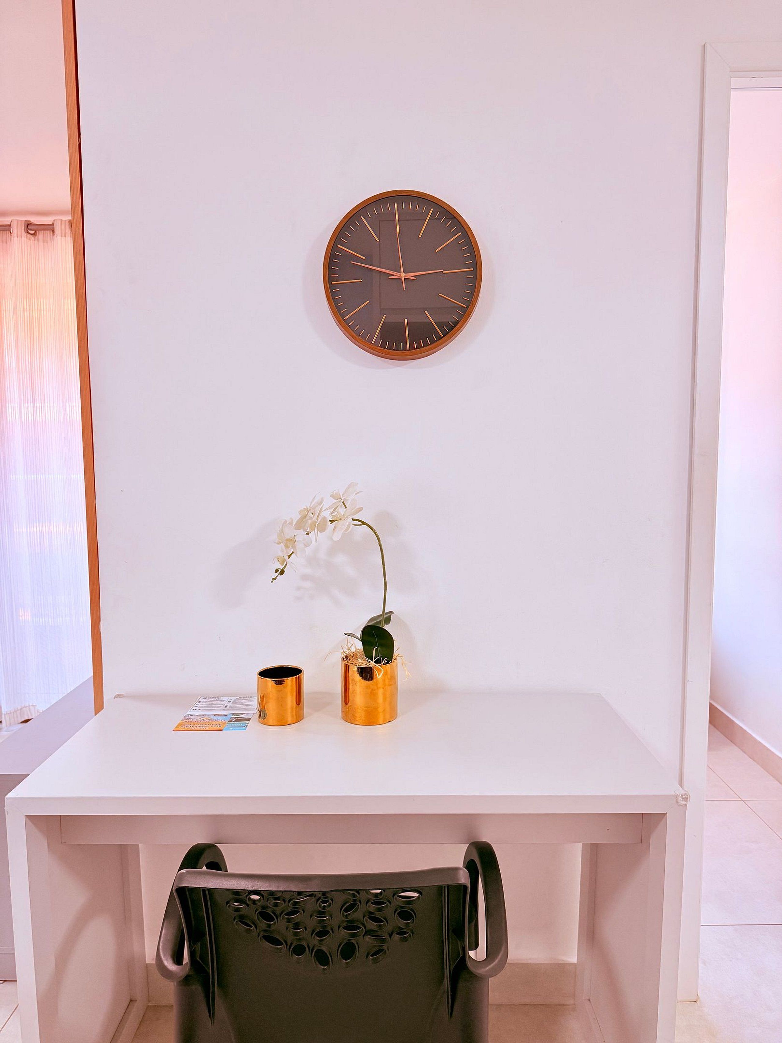 A white desk with a clock, two gold vases with plants, and a chair with a black backrest.