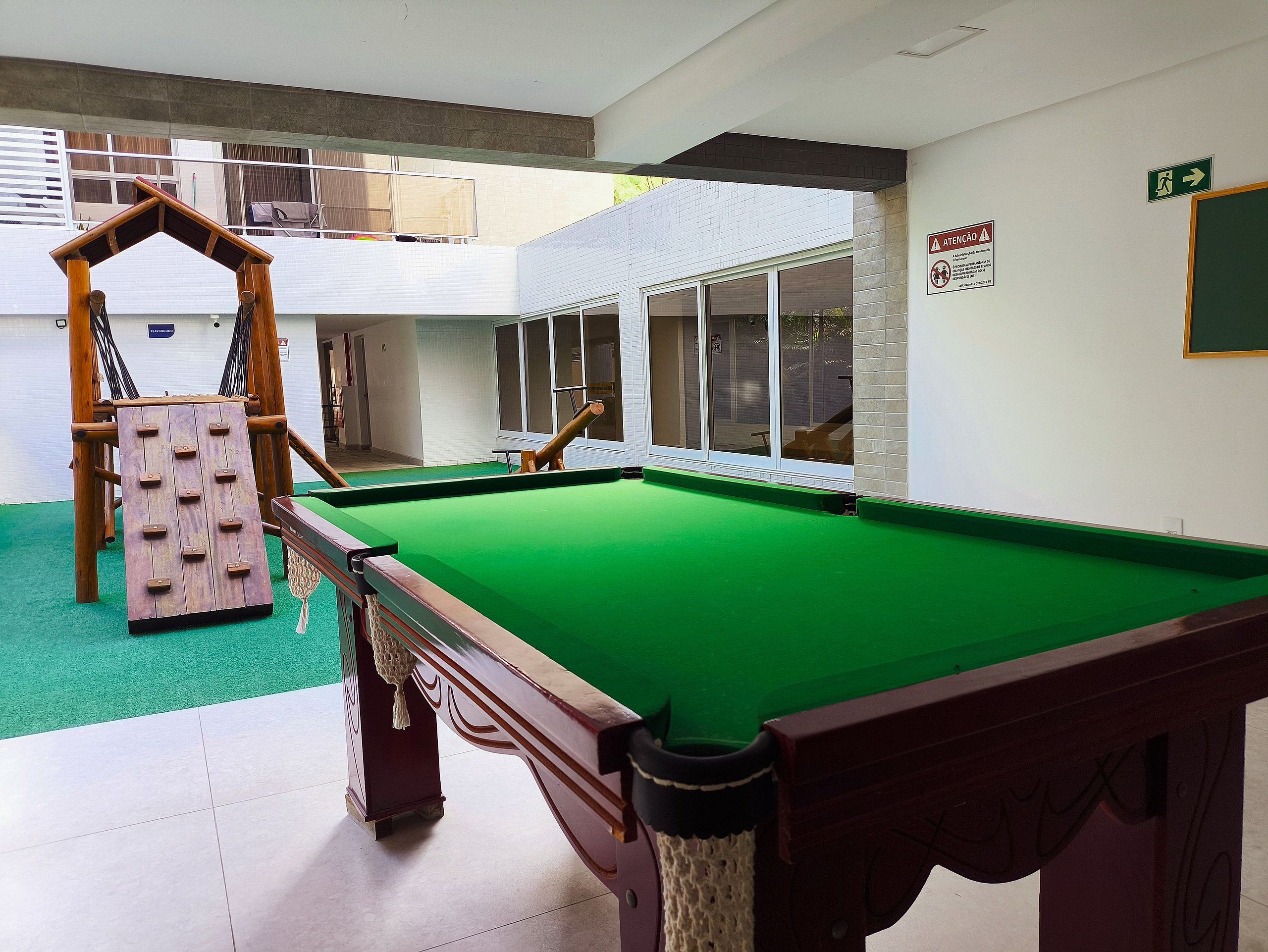 Una mesa de billar con superficie de fieltro verde, una estructura de juego de madera y vista a un edificio con amplias ventanas.