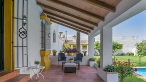 Un porche cubierto con mobiliario exterior, piscina y vista a un edificio tradicional.