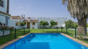 Una piscina con área cercada, un edificio blanco con techo de tejas y un árbol grande.