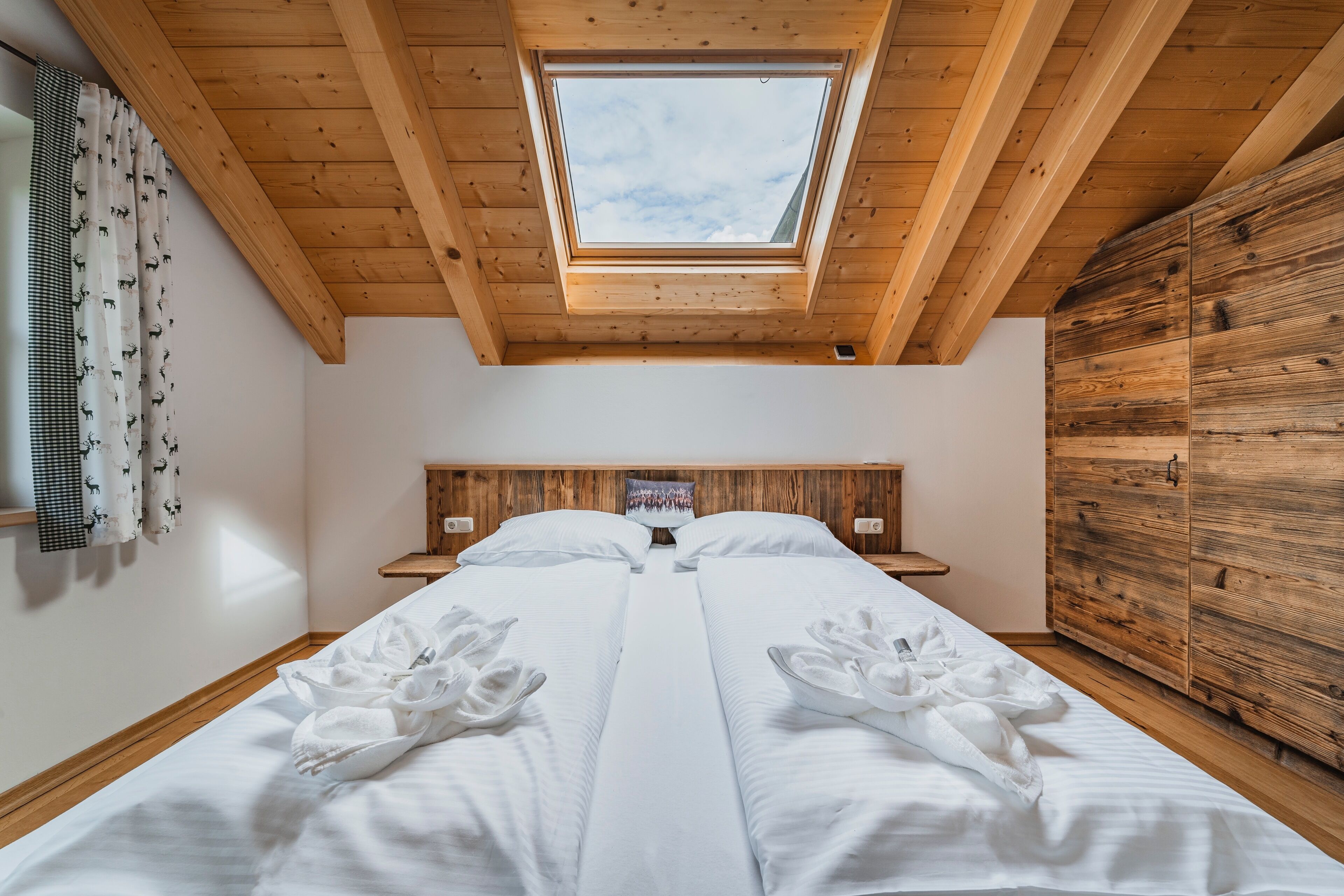 A bedroom with a skylight, wooden ceiling, two beds with white linens, a wooden wardrobe, and a headboard with a picture.