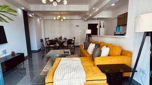 A modern living room with a yellow sofa, a coffee table, and a dining area in the background. - Exquisite 2-bedroom with 3 beds in Riverside, Nairobi (Nairobi)