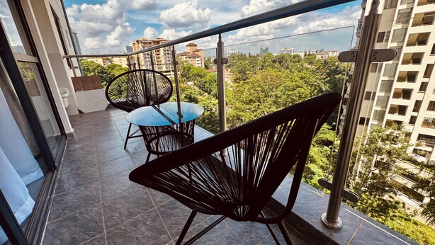 A balcony with a glass door, two black wicker chairs, a small round table, and a view of a cityscape with buildings and trees. - Exquisite 2-bedroom with 3 beds in Riverside, Nairobi (Nairobi)