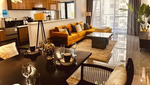 A modern living room with a sofa, coffee table, and dining table set up. There is a kitchen area with appliances and a window with curtains. - Exquisite 2-bedroom with 3 beds in Riverside, Nairobi (Nairobi)