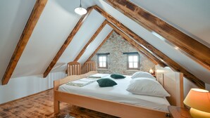 A bedroom with a sloped ceiling, wooden beams, a bed with pillows, a nightstand, and a lamp.