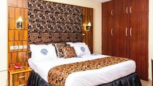 A bedroom with a patterned headboard, a bed with a black skirt, and wooden cabinets with two wall-mounted lamps. - Eagle Destiny Hotel and Apartment  (Asaba)