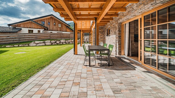 A covered patio area with a stone wall, wooden beams, and a table with chairs. - Dachstein West Apartment 1a (Russbach am Pass Gschuett)