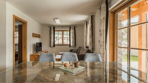 A modern dining area with a glass table, grey chairs, and a view of a wooden cabin through large windows. - Dachstein West Apartment 1a (Russbach am Pass Gschuett)