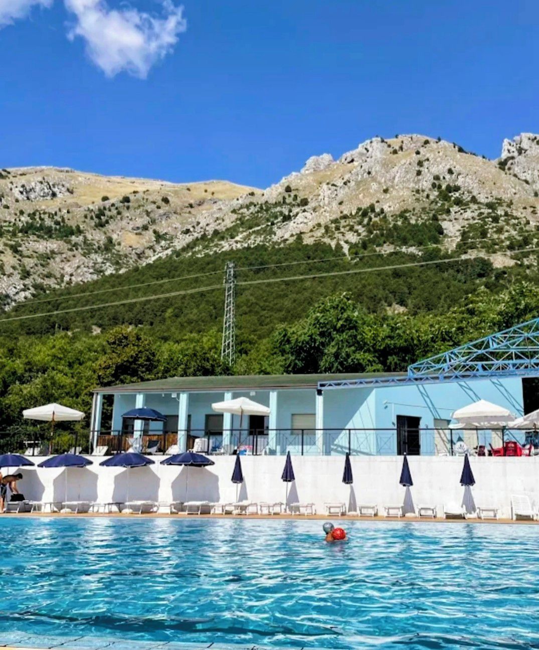 Ein Schwimmbecken mit Liegestühlen und Sonnenschirmen, ein Gebäude mit blauem Dach und eine gebirgige Kulisse.