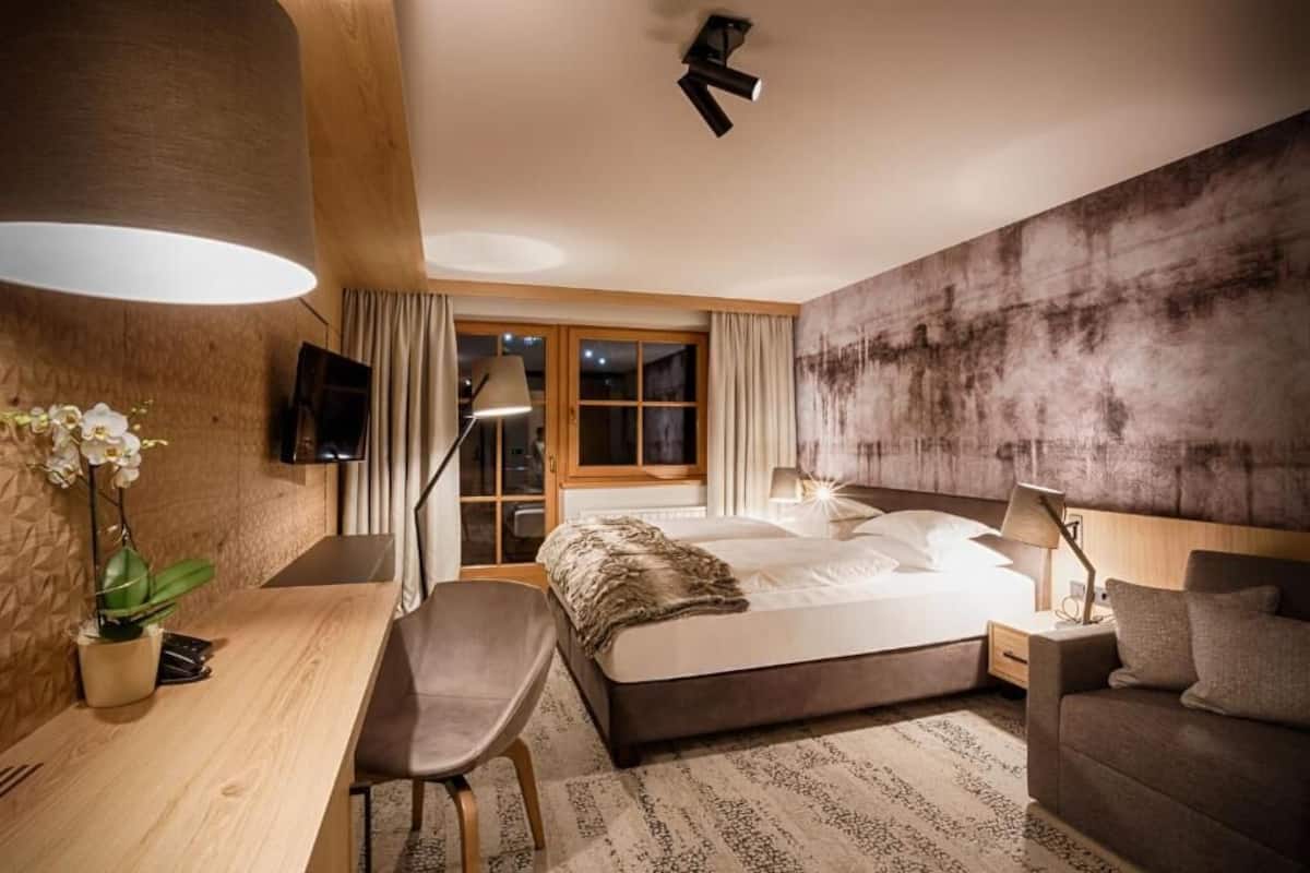 A modern hotel room with a large bed, a desk, a chair, a TV, and a large window with curtains.